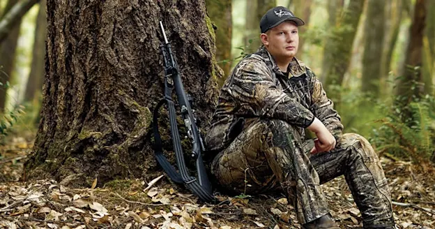 A young hunter in full camo gear sits by a tree in the woods.