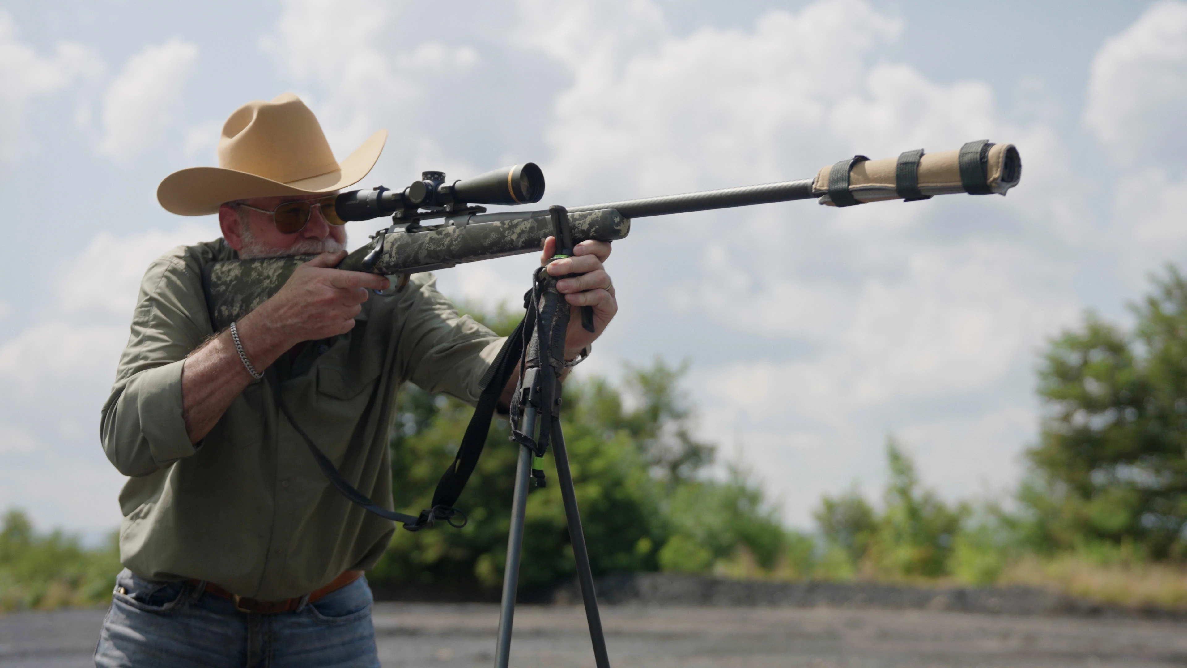 A shooter in a straw hat fires a suppressed rifle in shooting sticks.