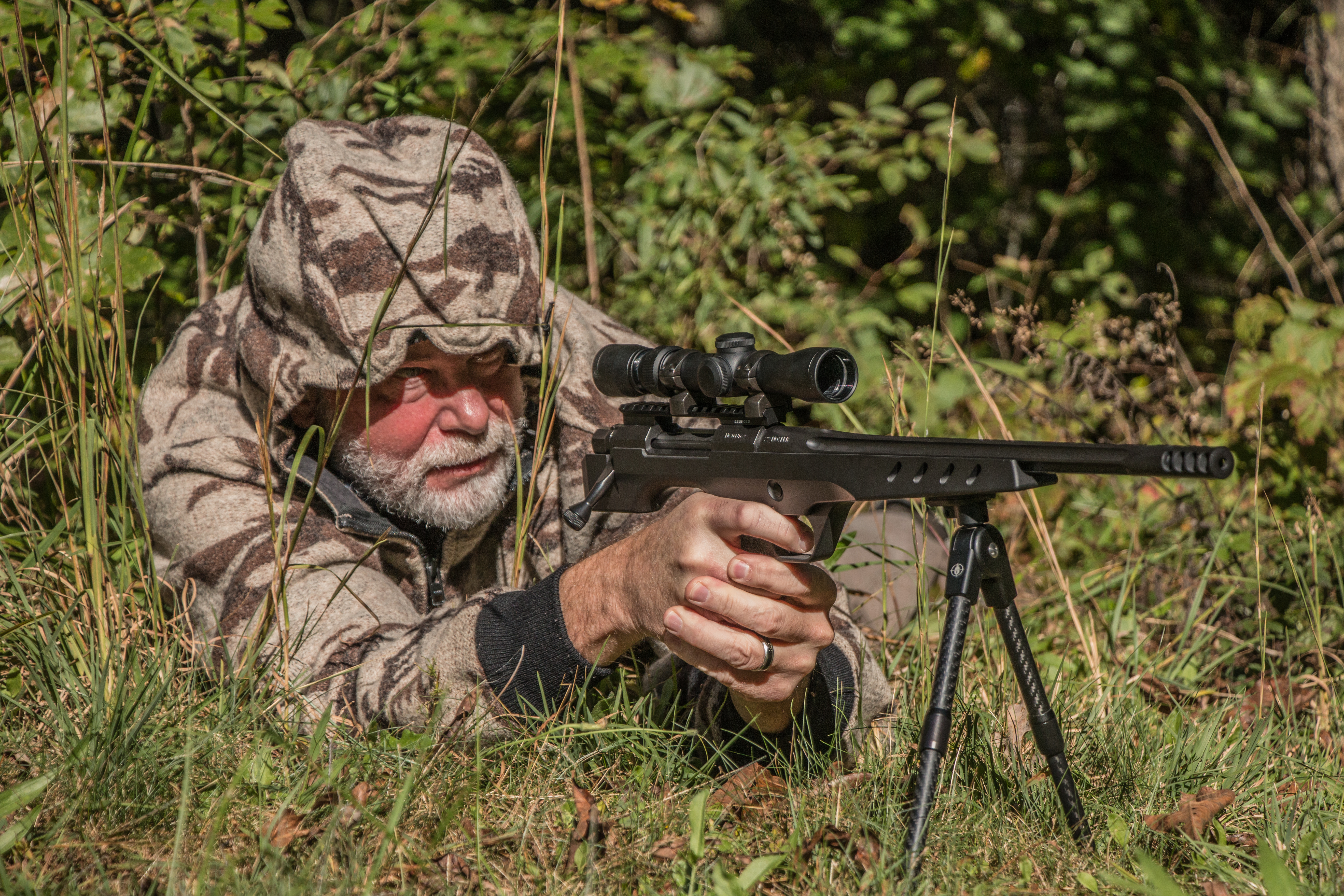 A handgun hunter hidden in grass takes aim trough a scope. 