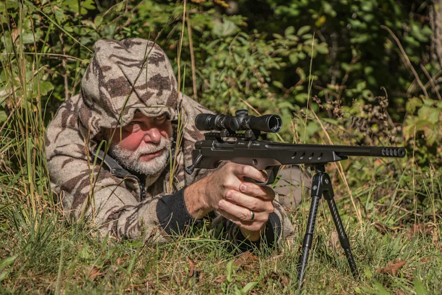 A handgun hunter hidden in grass takes aim trough a scope.