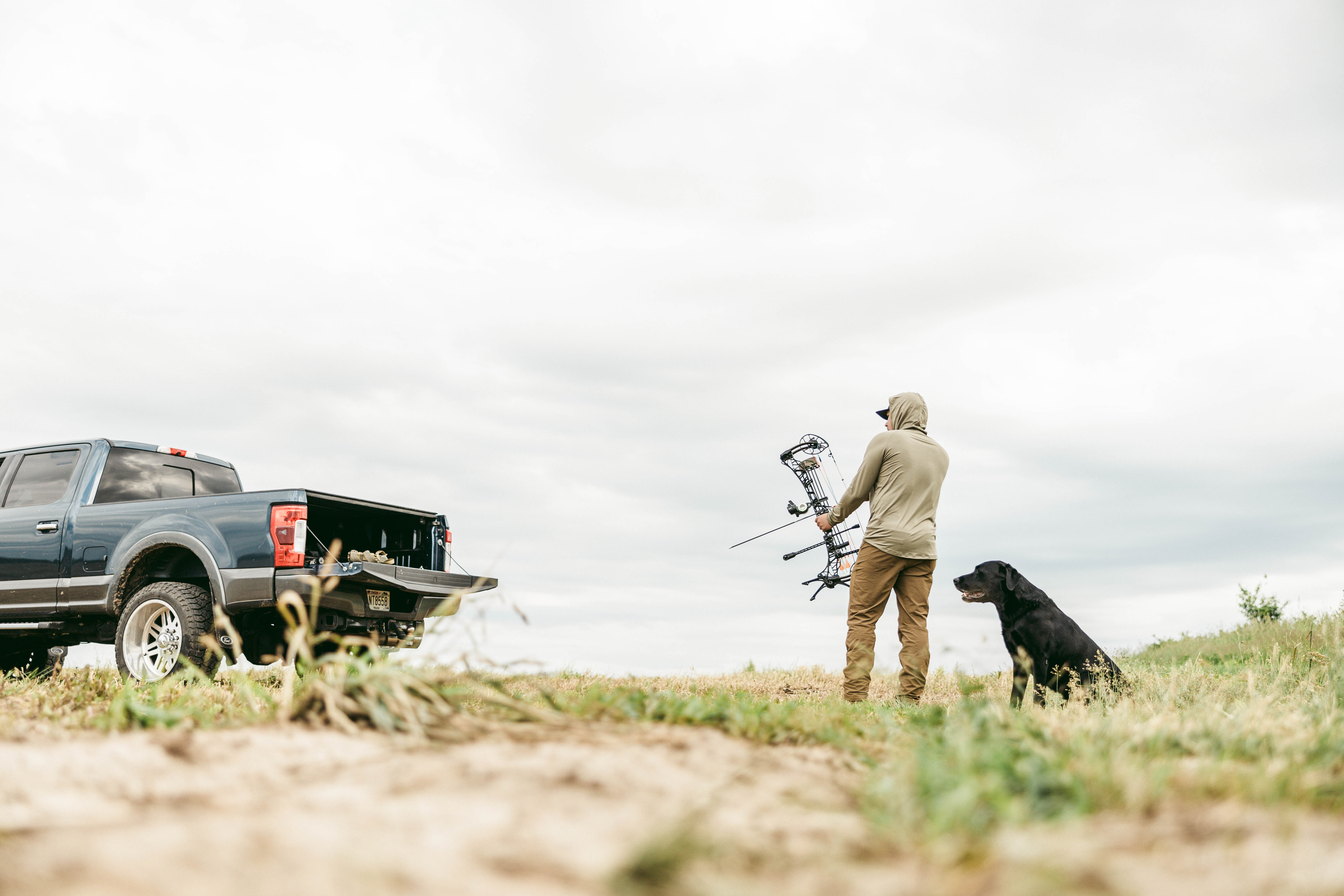 A bowhunter shoots at a target in a field with his dog standing behind him. 