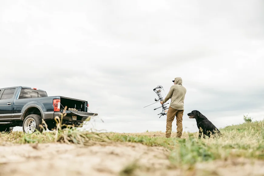 A bowhunter shoots at a target in a field with his dog standing behind him.