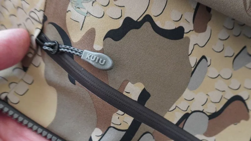 A close-up of a zipper and pull on a black, brown, and tan Kuiu Yukon TR rain jacket with a hand partially visible. 