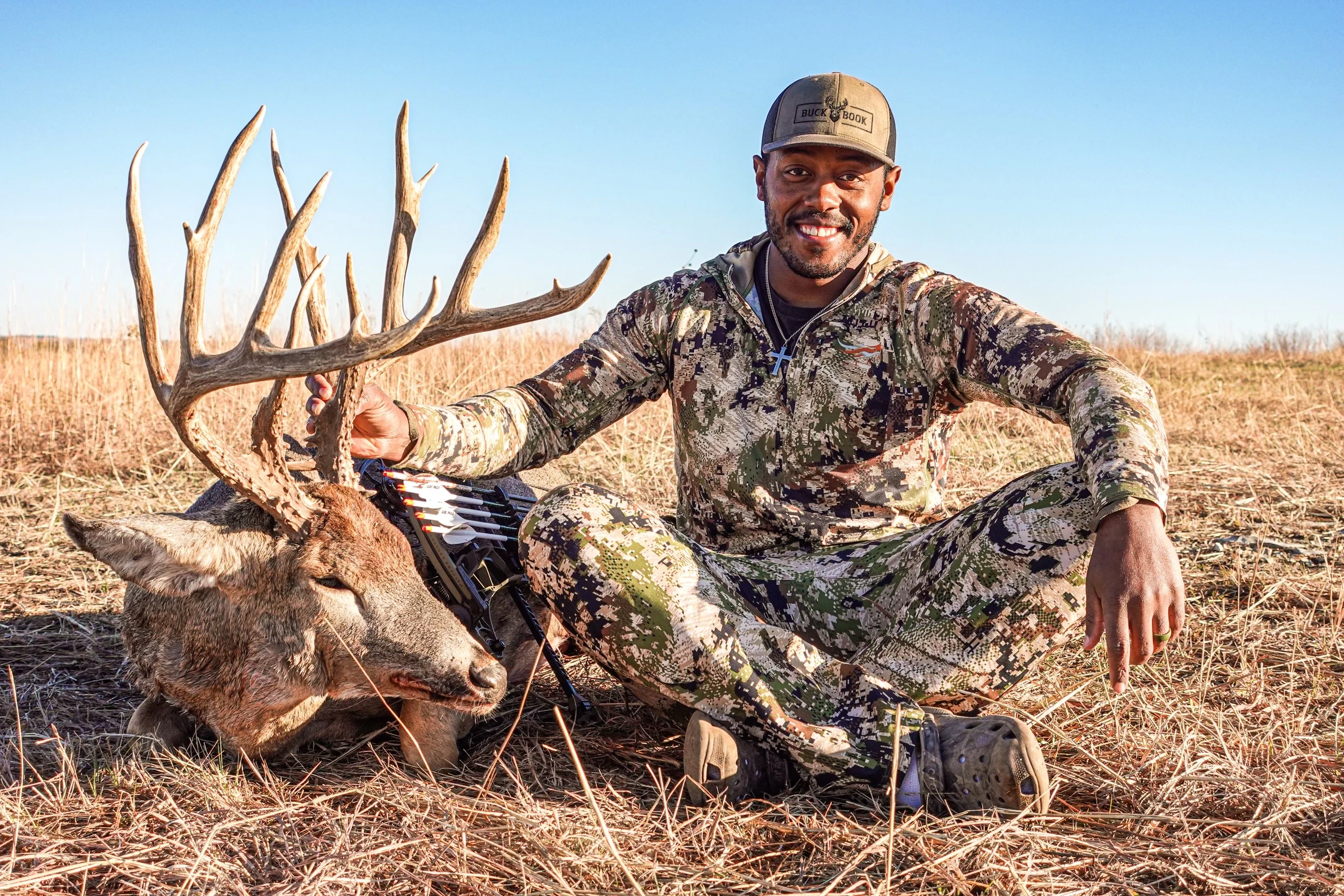 A hunter sits of the ground and poses with a huge whitetail buck he took with a bow. 