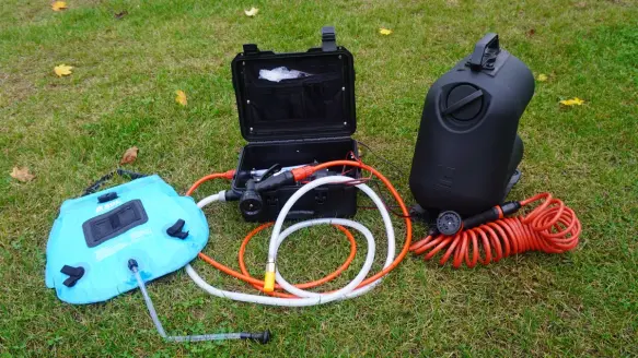 Three camping showers of different designs, one blue and two black, sitting on a green lawn.