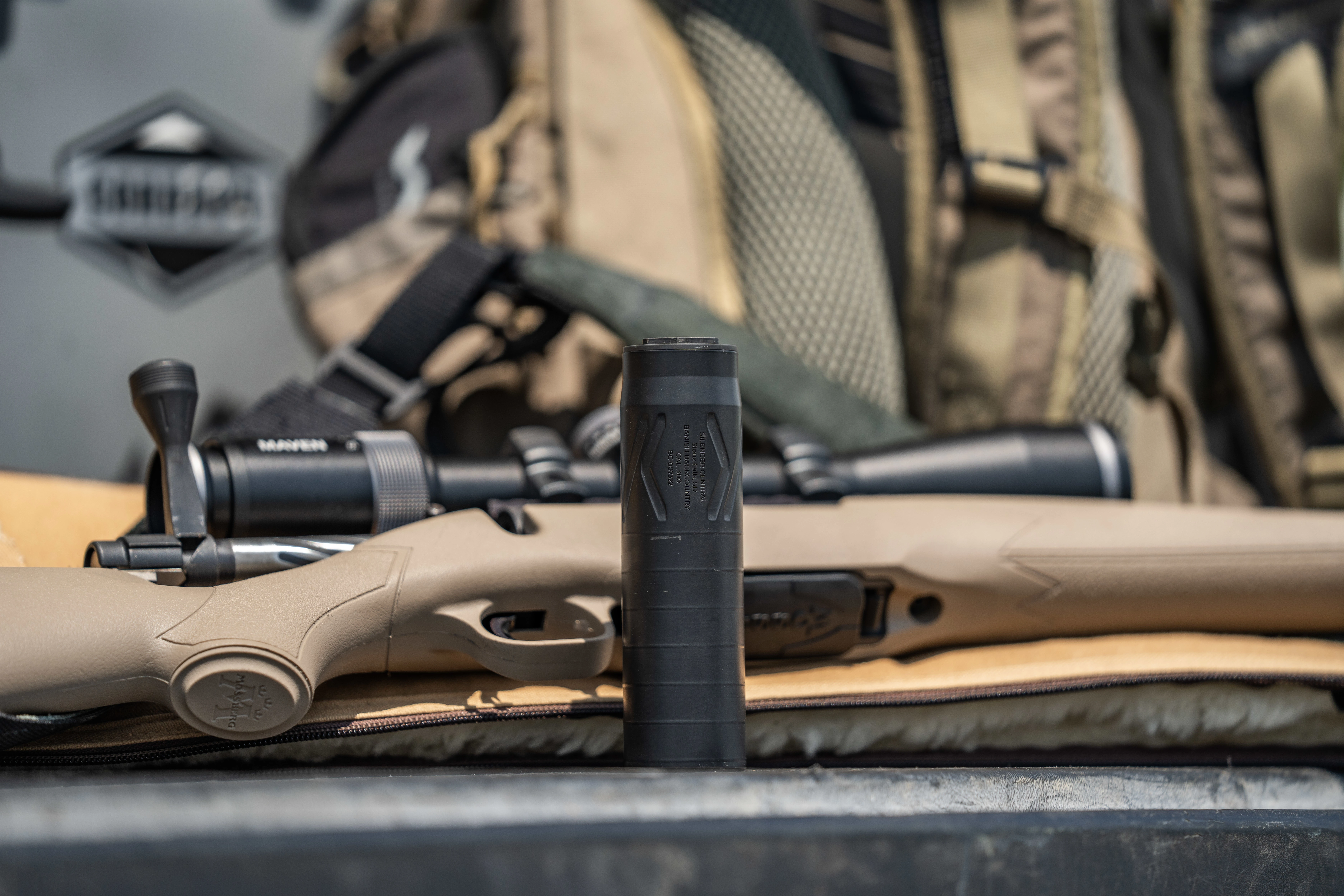 A suppressor sits upright next to a rifle on a truck bed