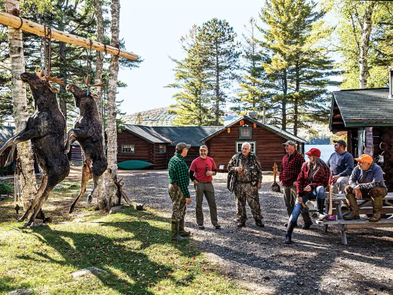 A group of hunter stand outside a hunting cabin with two moose hanging from a pole.