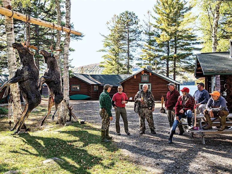 A group of hunter stand outside a hunting cabin with two moose hanging from a pole.