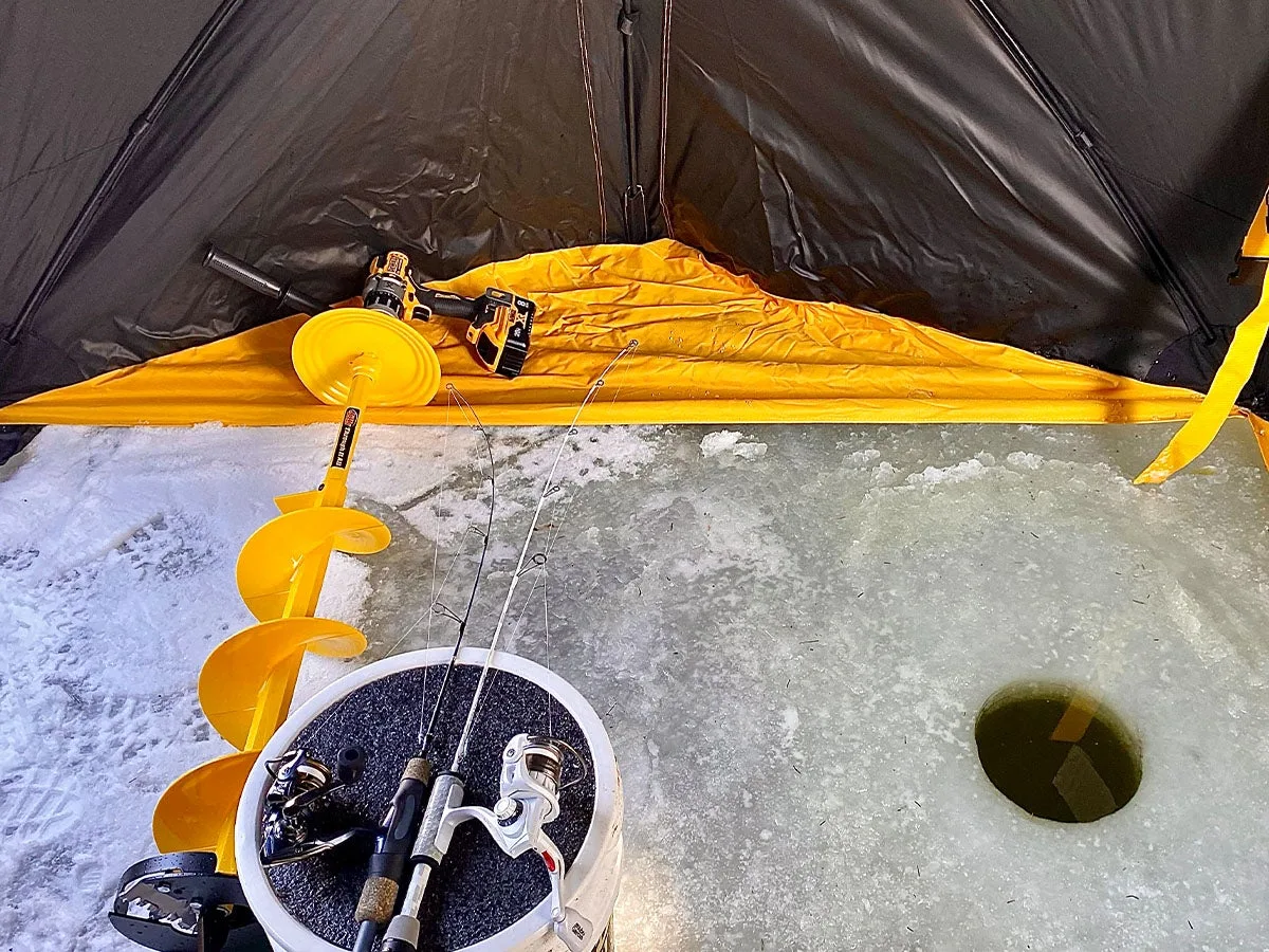 An ice auger, drill fishing poles, and bucket next to a hole int eh ice insdie a shanty