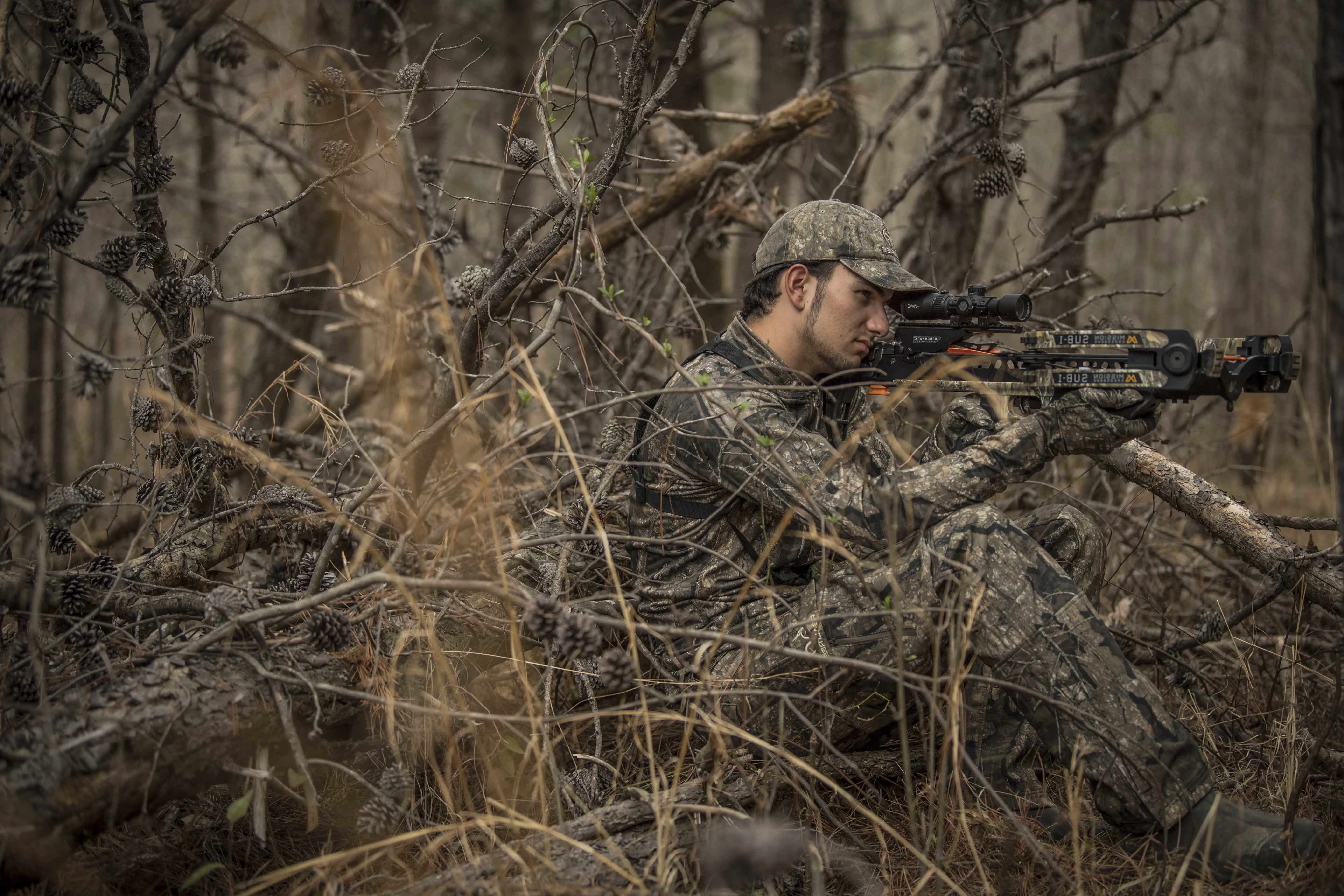 A hunter shoots a crossbow from a natural blind with open woods in the background. 