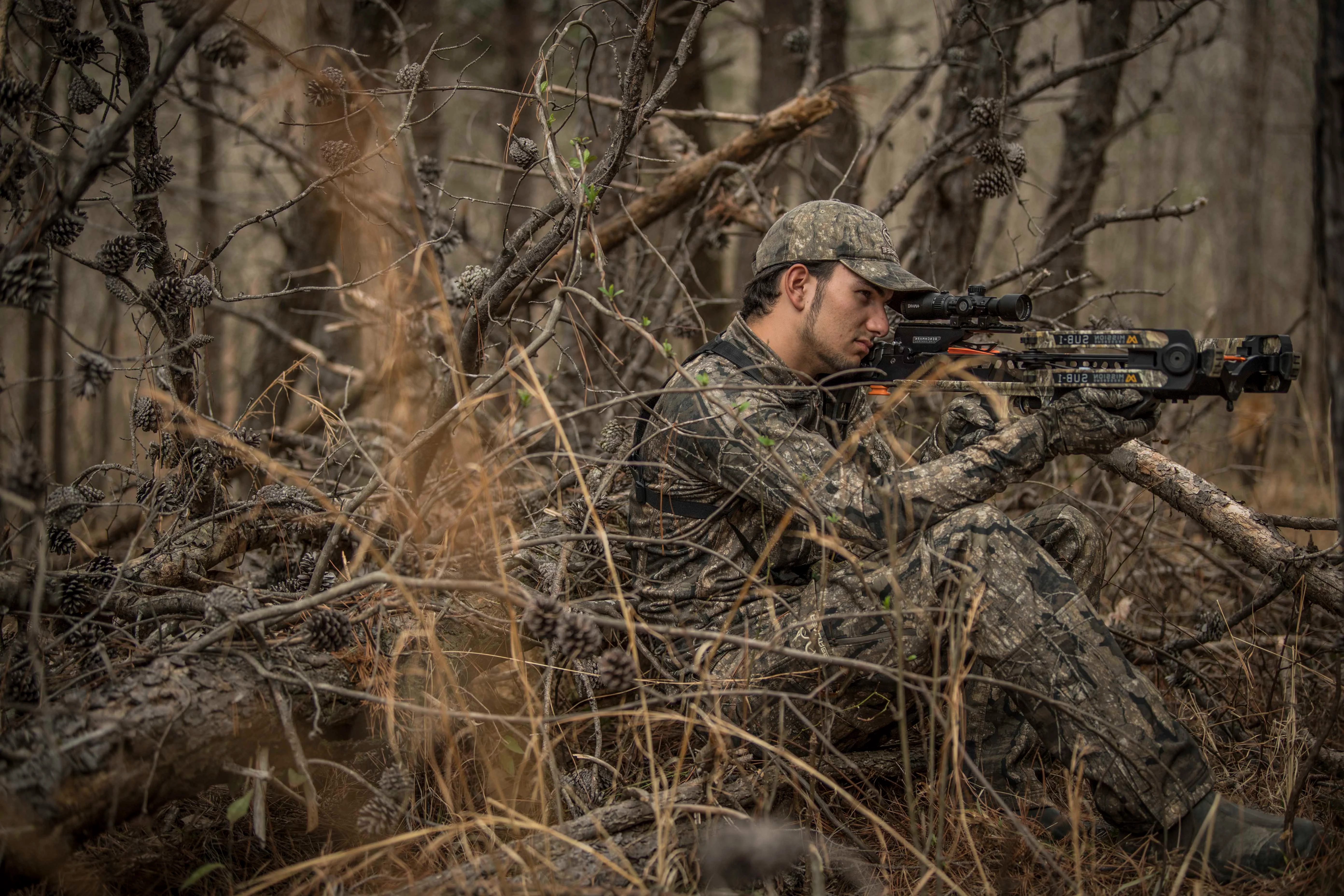 A hunter shoots a crossbow from a natural blind with open woods in the background. 