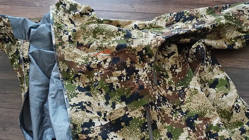 A green, tan, and black Sitka Dew Point jacket sitting on a hardwood floor with the gray inner lining visible. 