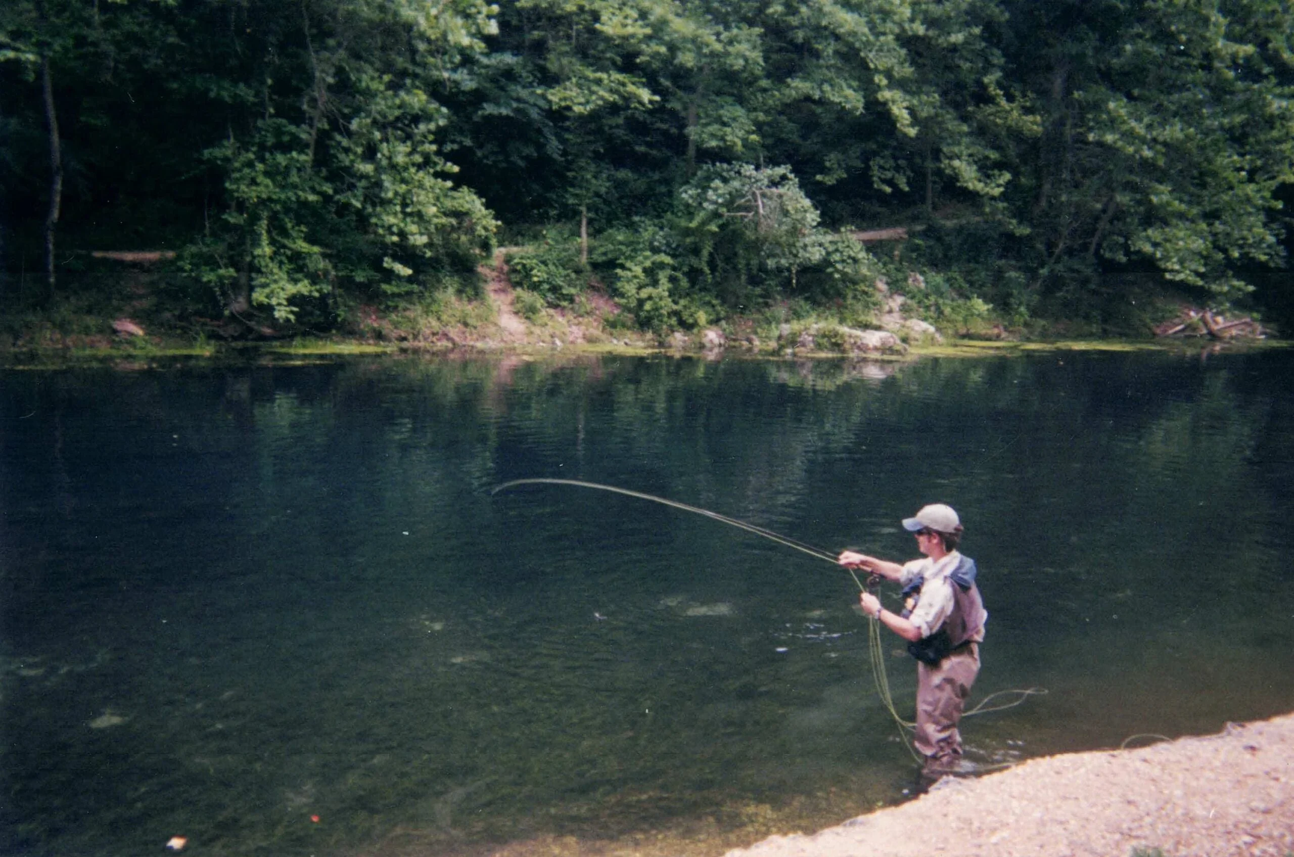 A fly fisherman stands at the edge of a trout stream holding a fishing rod.