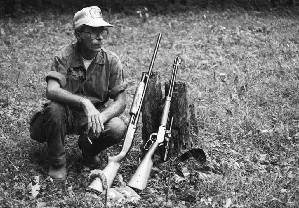 Squirrel hunter with a shotgun and .22LR rifle sitting on the ground.