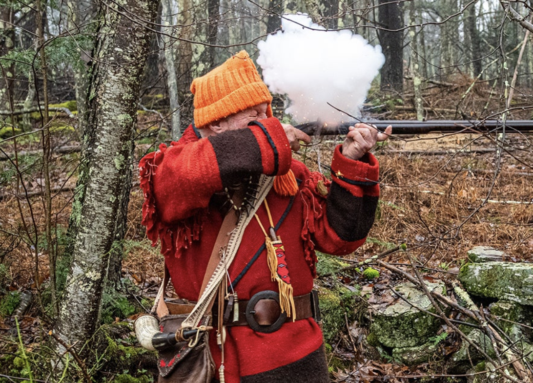 A man dressed in traditional garb fires a flintlock rifle. 