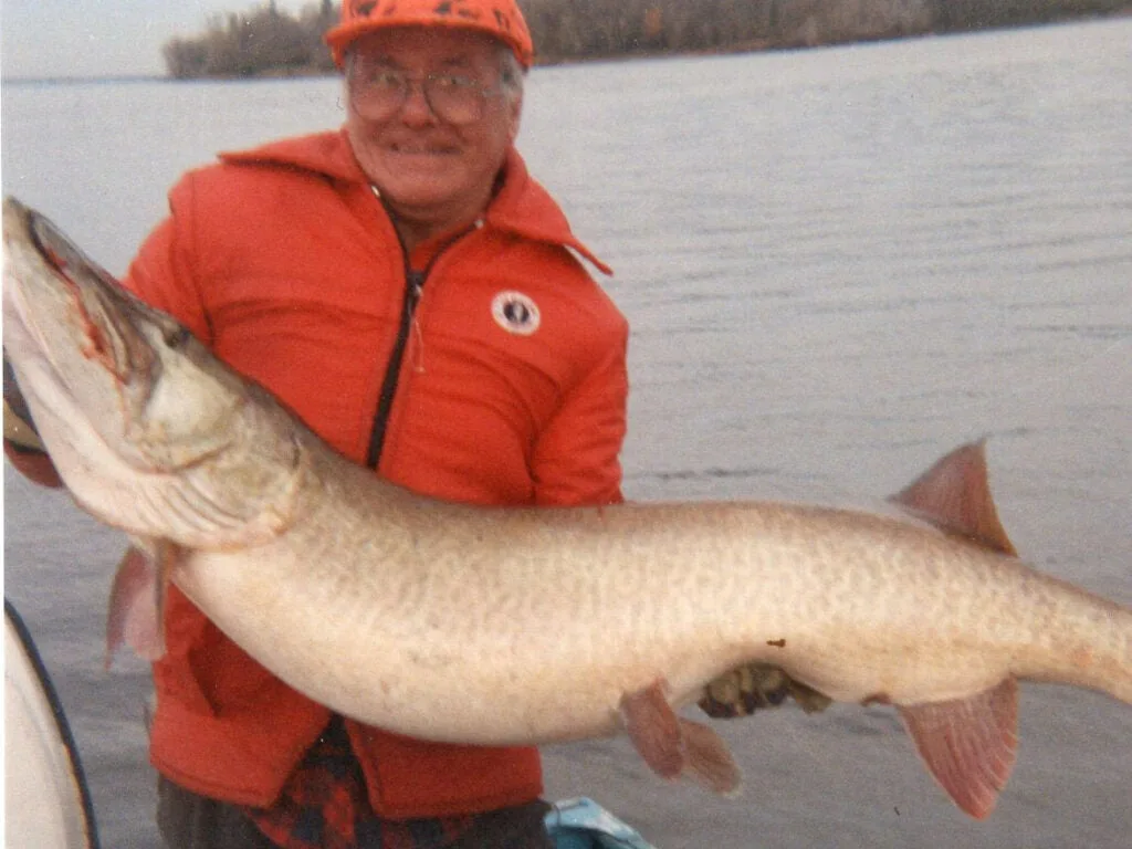 A man in an orange vest holds up a large muskie.