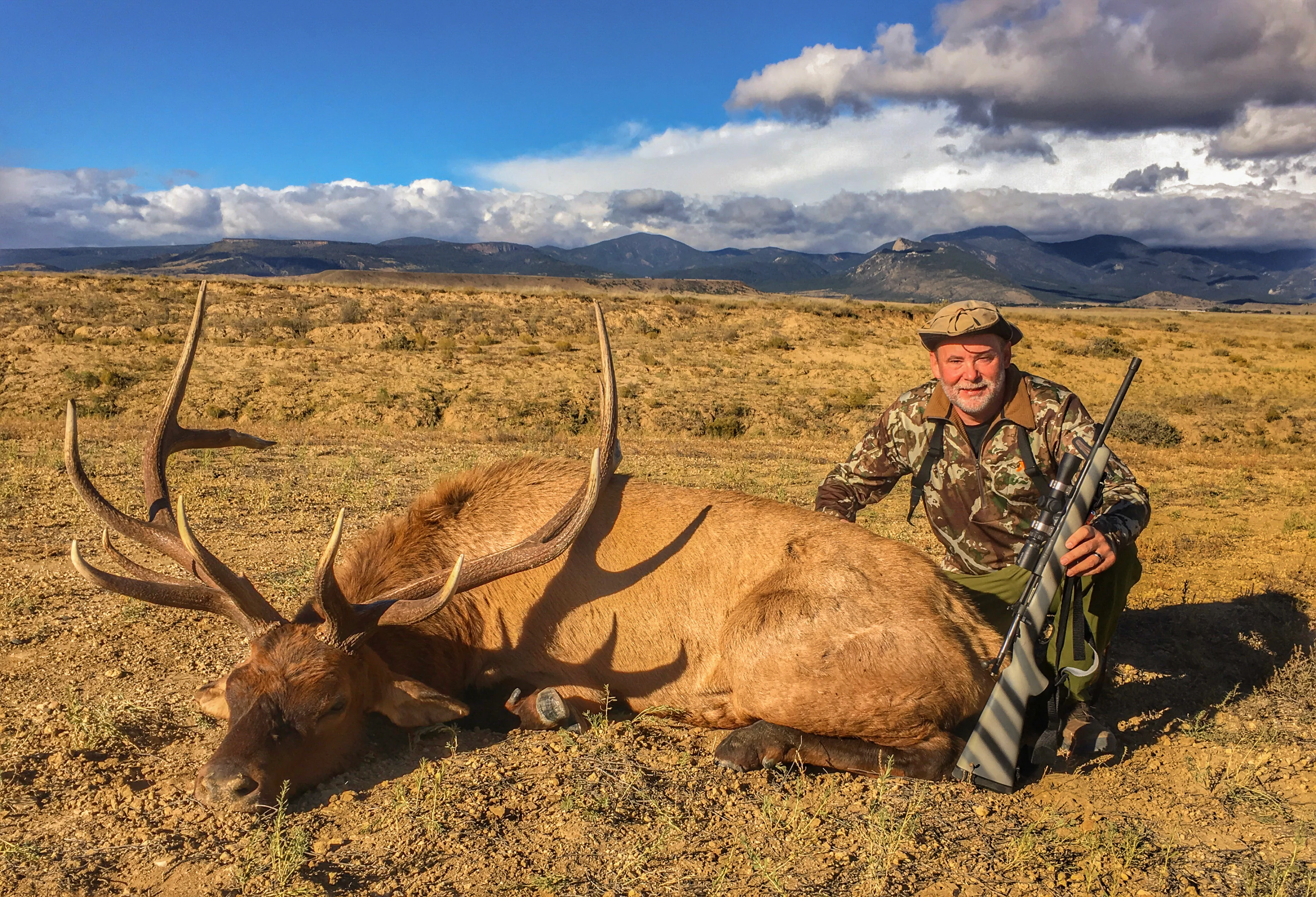 A poses with a nice bull elk taken with a rifle with mountains in background. 