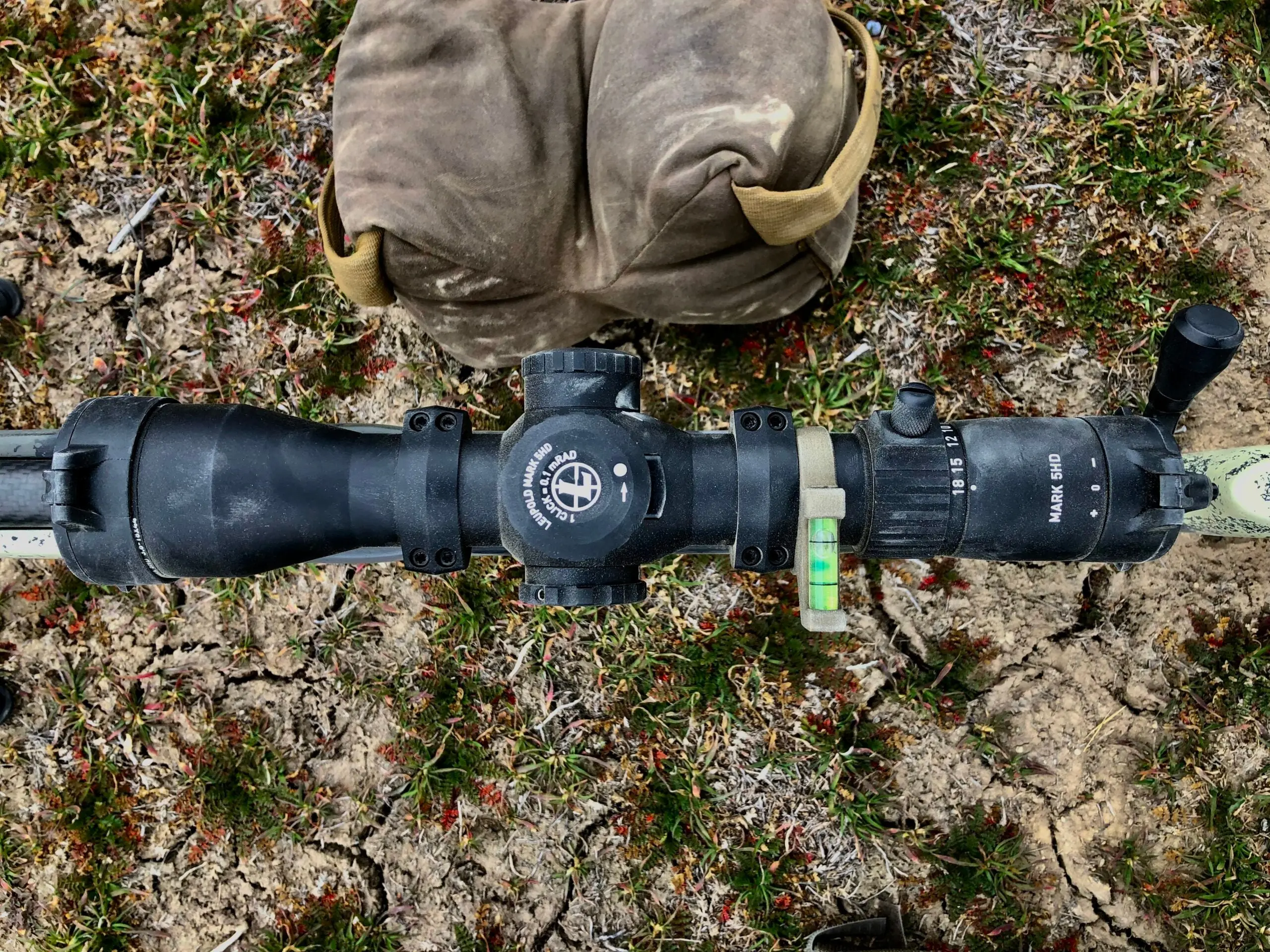 A long-range rifle scope with MRAD turrets, as opposed to MOA, sitting on the ground near a sand bag.