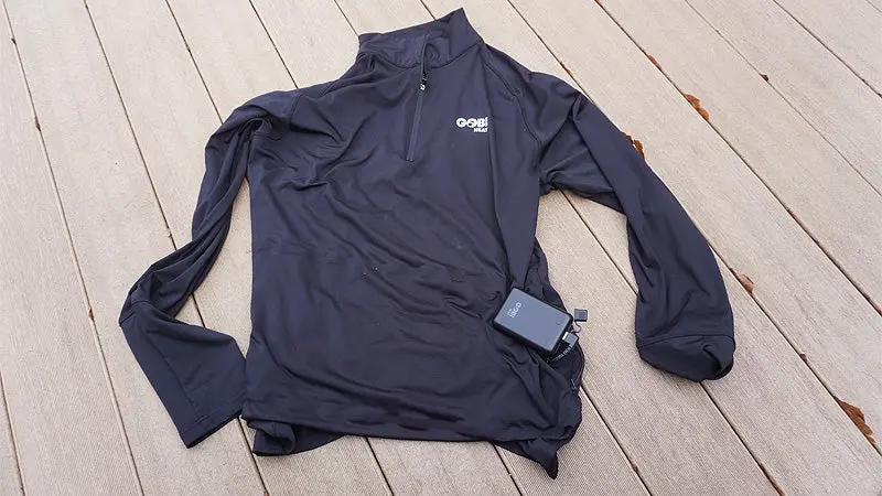 A black Gobi Heat Basecamp base layer with a battery bank outside the pocket on a tan deck. 