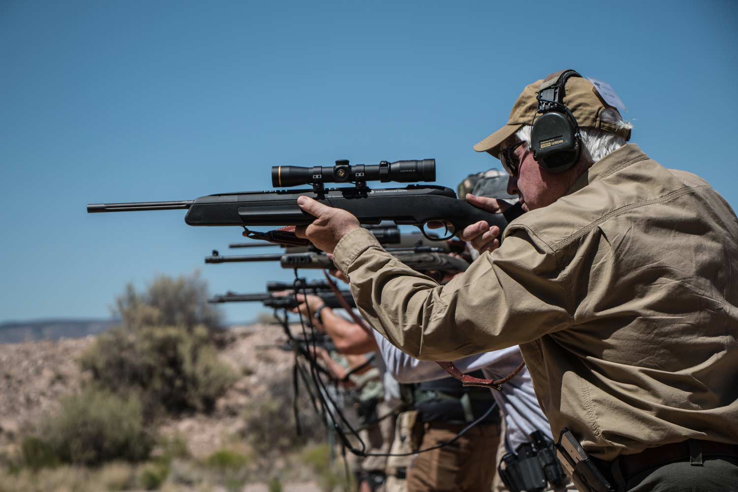 A line of men aim scout rifles at a shooting range.