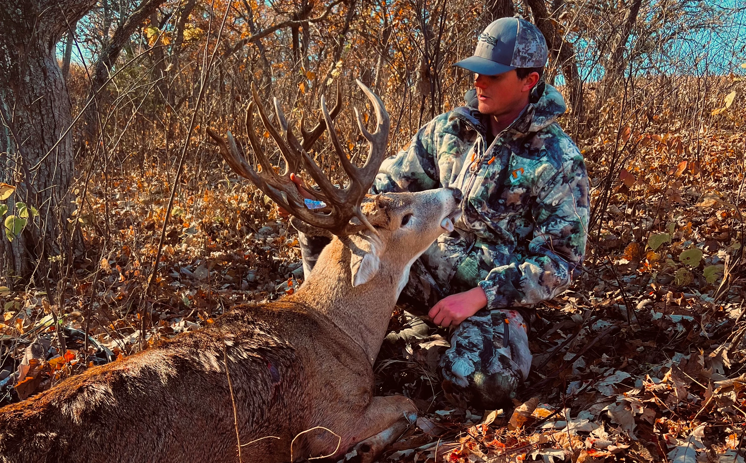 A Kansas hunter sits on the ground and poses with a trophy buck he took with a bow. 