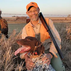 A hunter dressed in tan and orange hunting close knees beside a german shorthaired pointer
