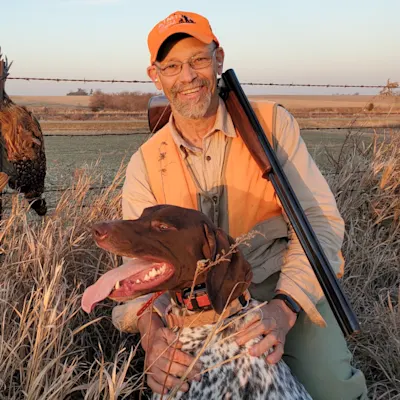 A hunter dressed in tan and orange hunting close knees beside a german shorthaired pointer