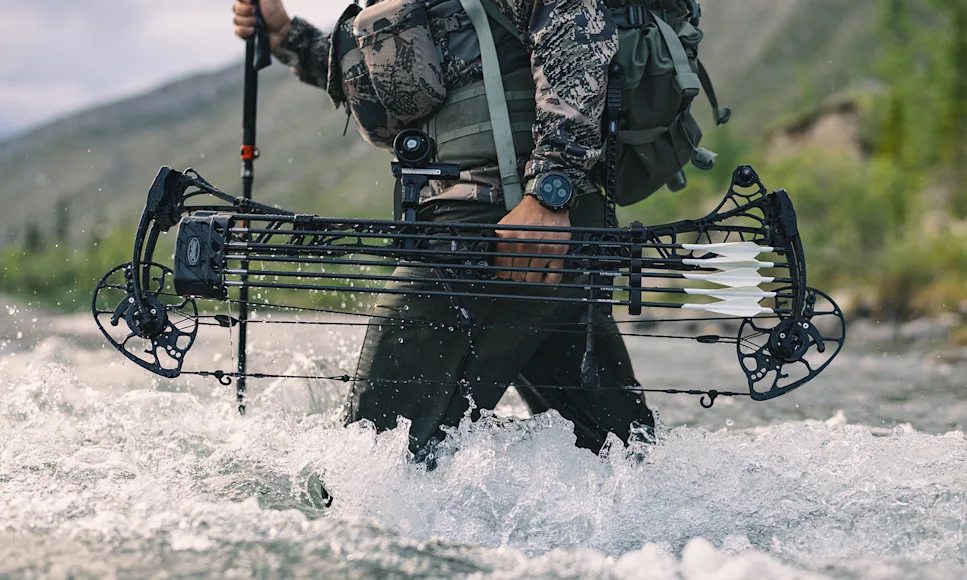 A hunter carries a compound bow as he fords a creek.
