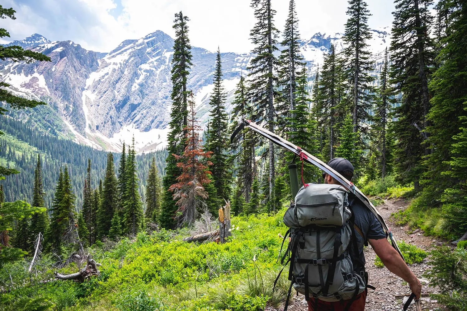 hiker carries crosscut saw down trail, snowy mountains in distance across valley