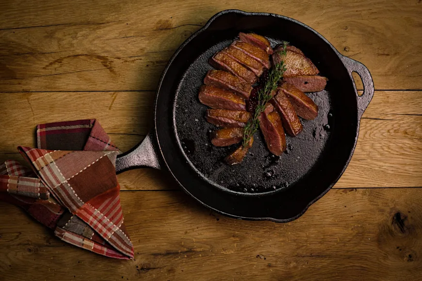 Sliced duck breast in a black cast iron skillet resting on a wooden table.