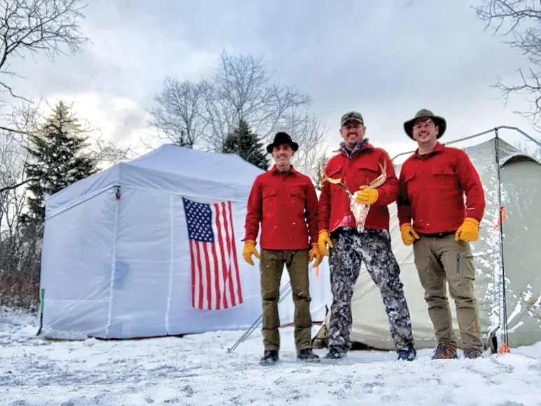 Three men in red stand in the snow holding a deer head skull.