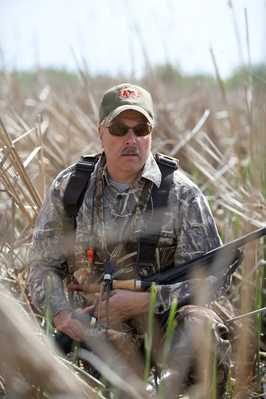 Duck hunter sits in tall reeds while holding a shotgun