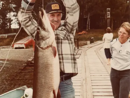 An older man holds up a large muskie while a lady looks on.