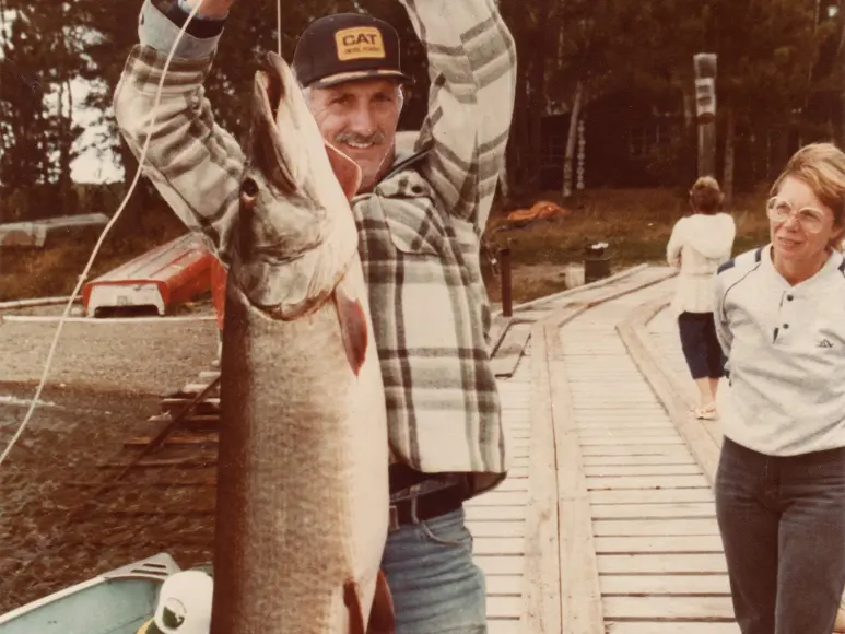 An older man holds up a large muskie while a lady looks on.