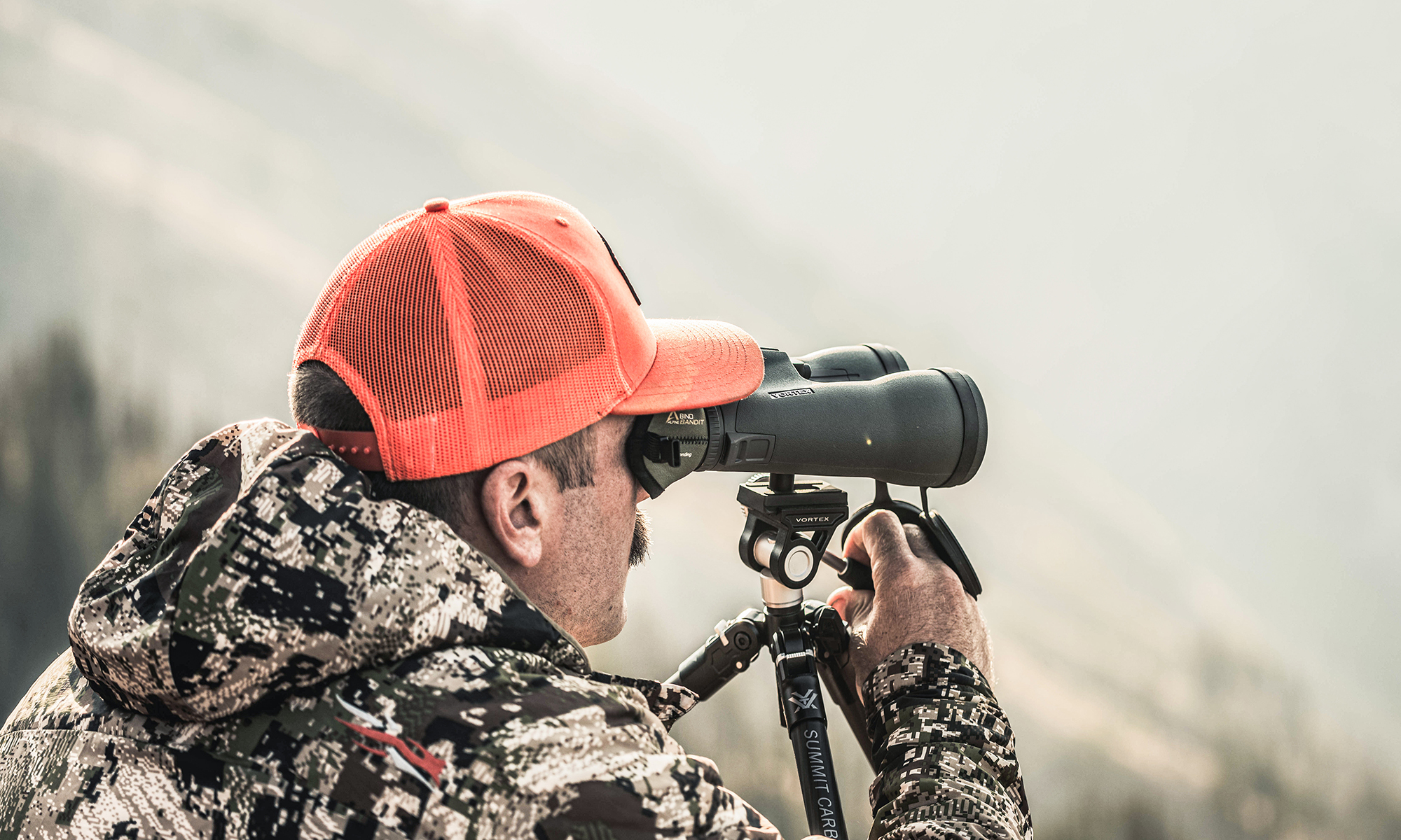 A hunter uses binoculars in a mountain setting.
