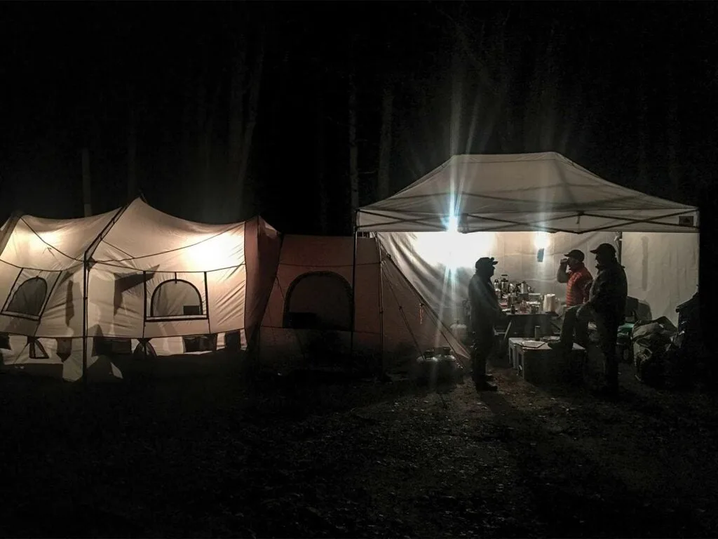 Groups of hunters stand outside of a lit camping tent at night.