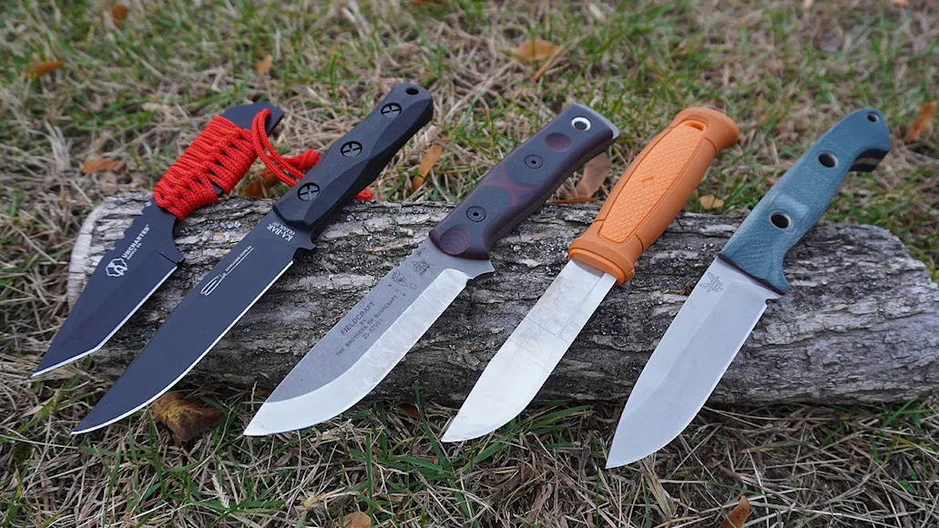 Five fixed blade bushcraft ready knives sitting on a log on a grassy lawn.