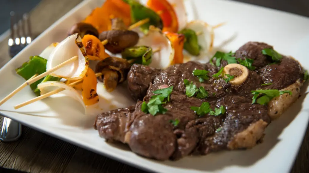 Bear steak on a white plate with vegetable kabobs.