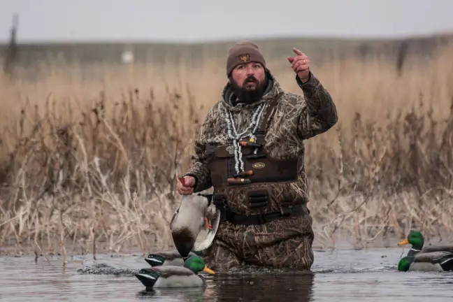 Duck hunter standing in water holding a duck and pointing towards the sky