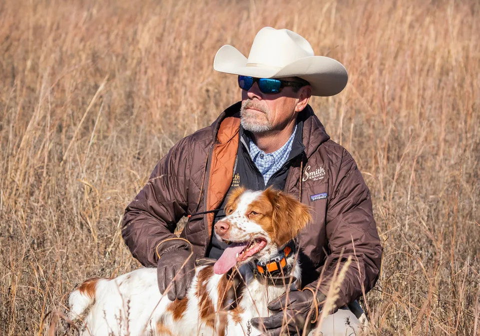 A dog trainer with a white cowboy hat and brown jacket works with a Brittany spaniel in a weedy field. 