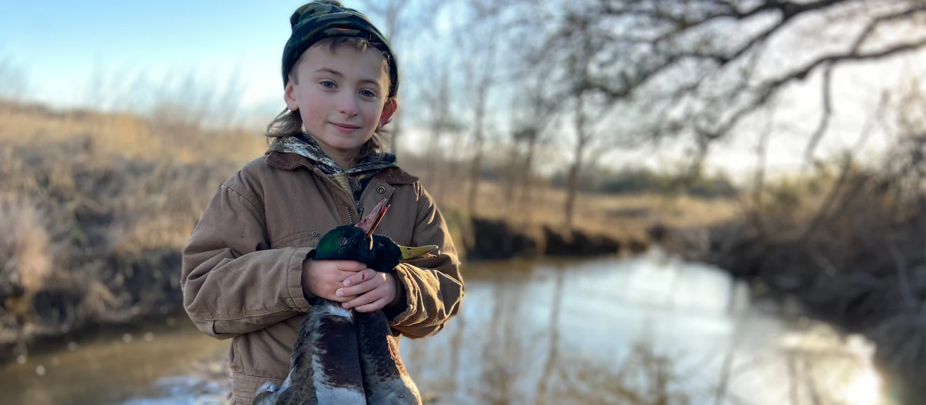 boy holds two ducks and stands in front of pond in lightly wooded area