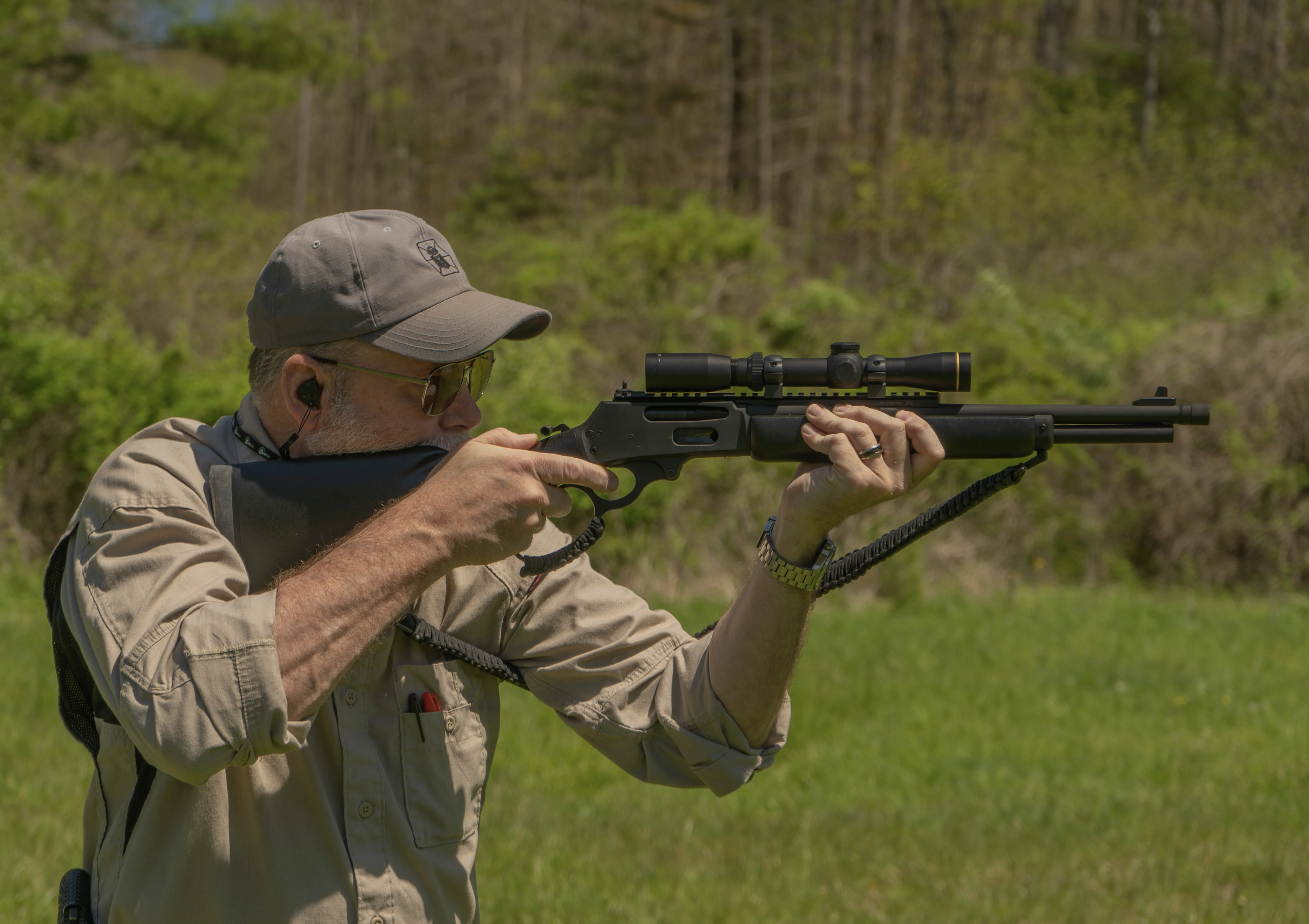 A shooter fires a scoped lever-action rifle from the off hand position, with woods in background. 