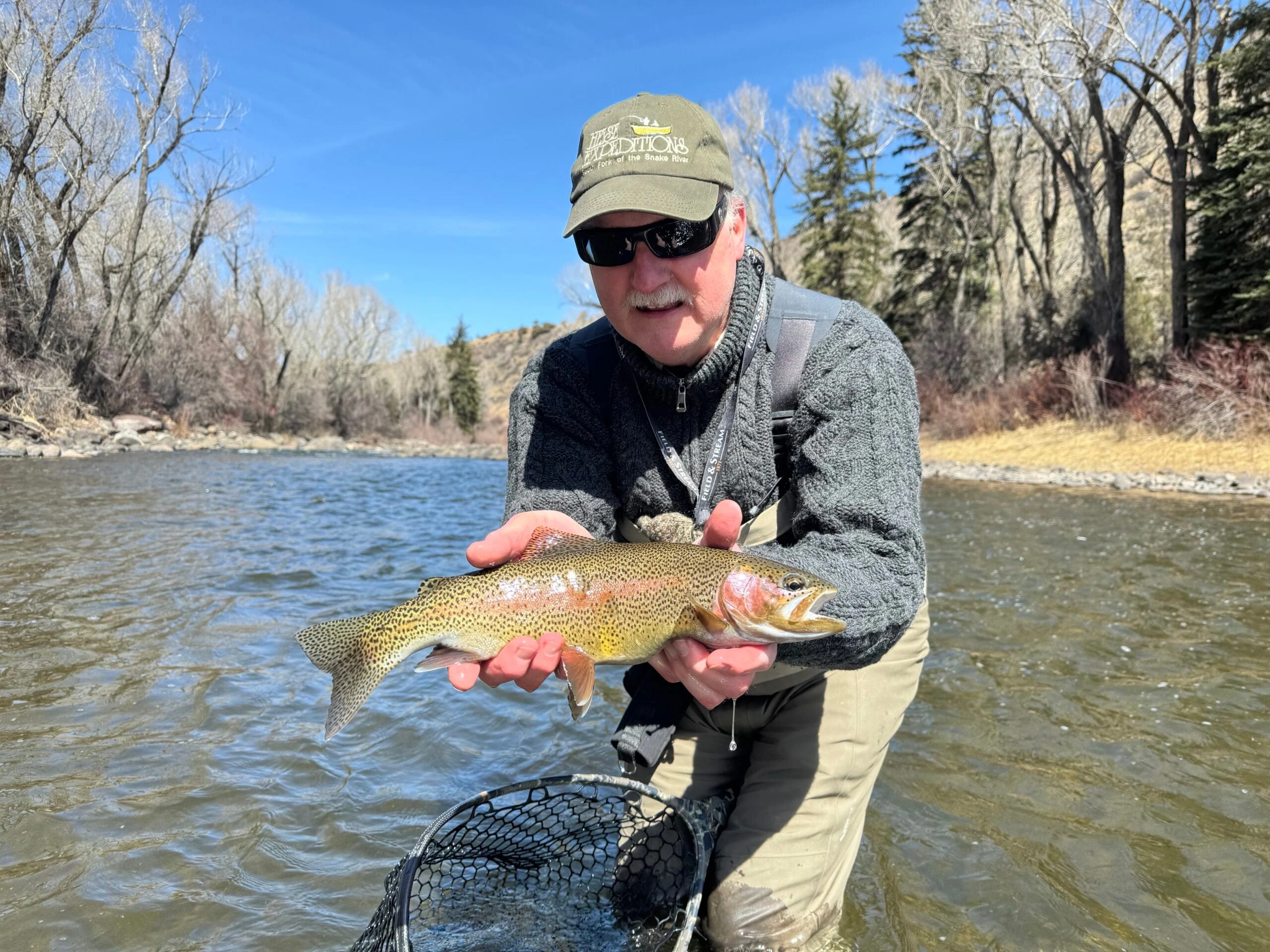 A fisherman stands in a river lined with trees holding a rainbow trout.