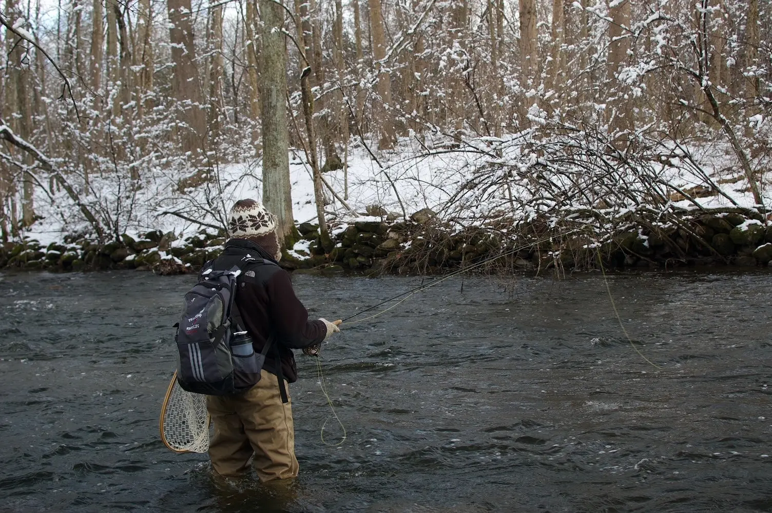 Fly fisherman stand in a river while making a cast with snow covered banks in the backgrond