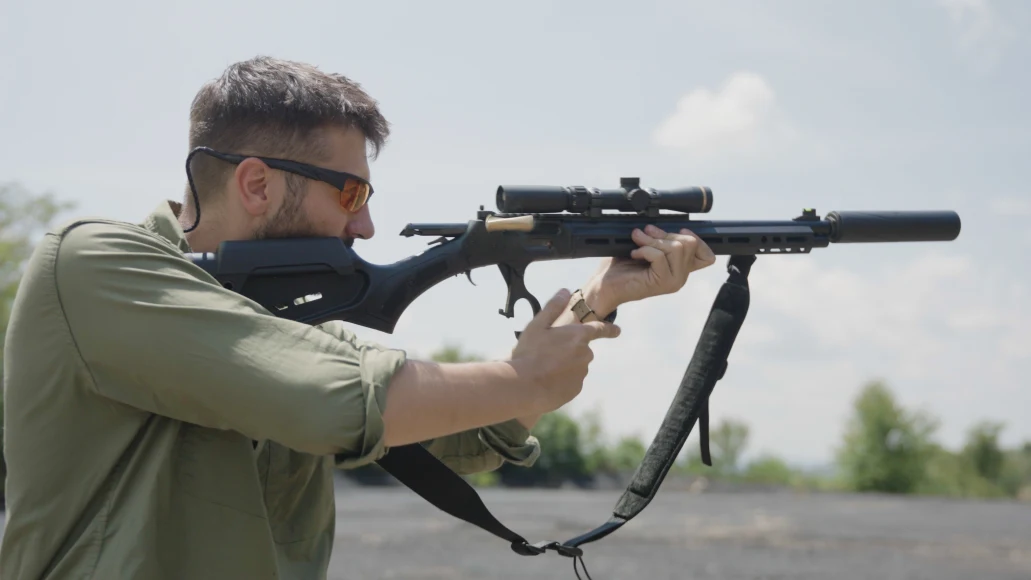 A man in a green shirt and shooting glasses fires a rifle with a suppressor on it.