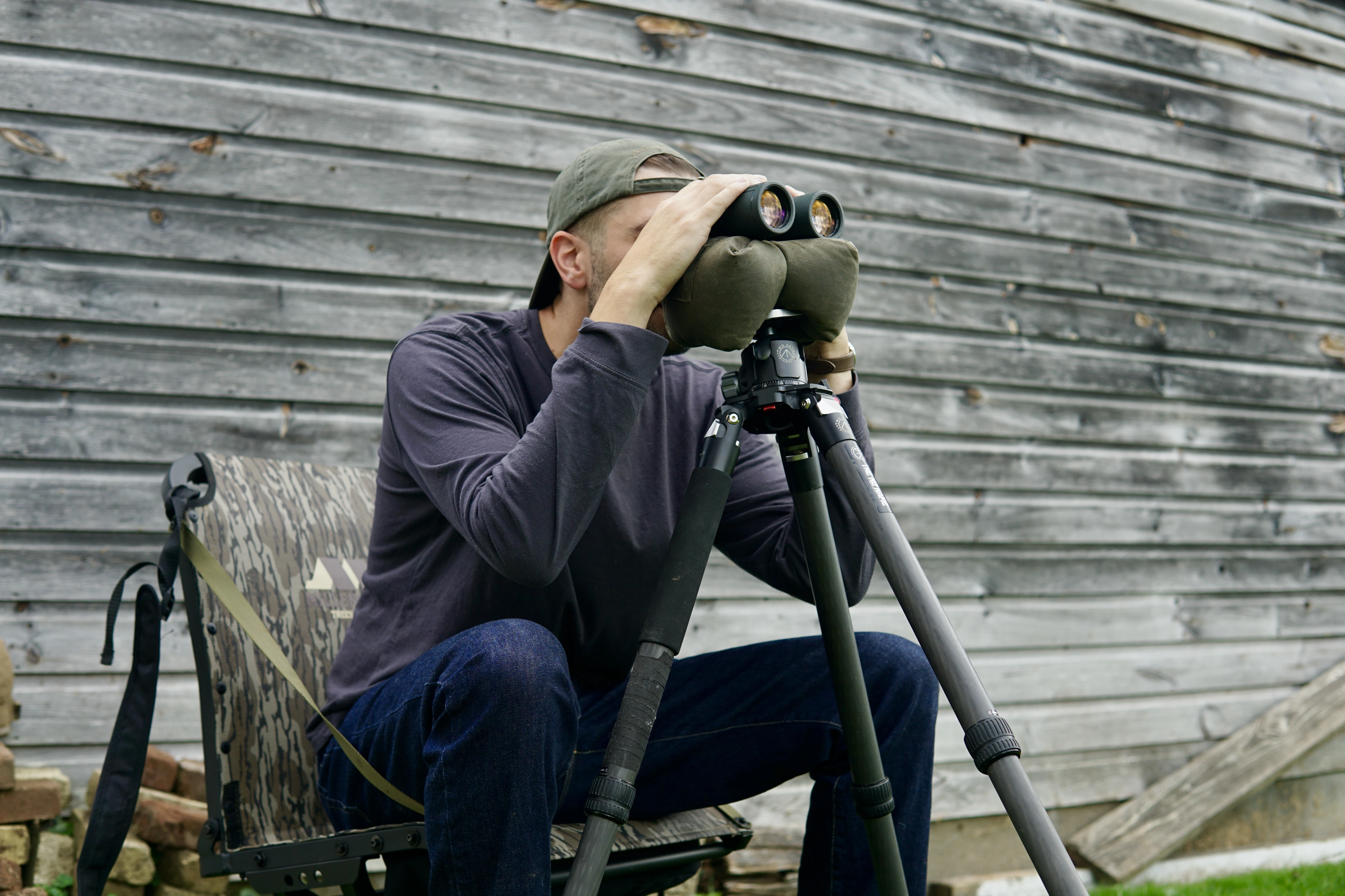 A man looks through the Hawke Frontier LRF binoculars from a chair in front of a barn wall.