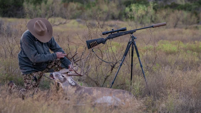 A hunter admires a whitetail buck taken with the rifle sitting on a tripod nearby. 