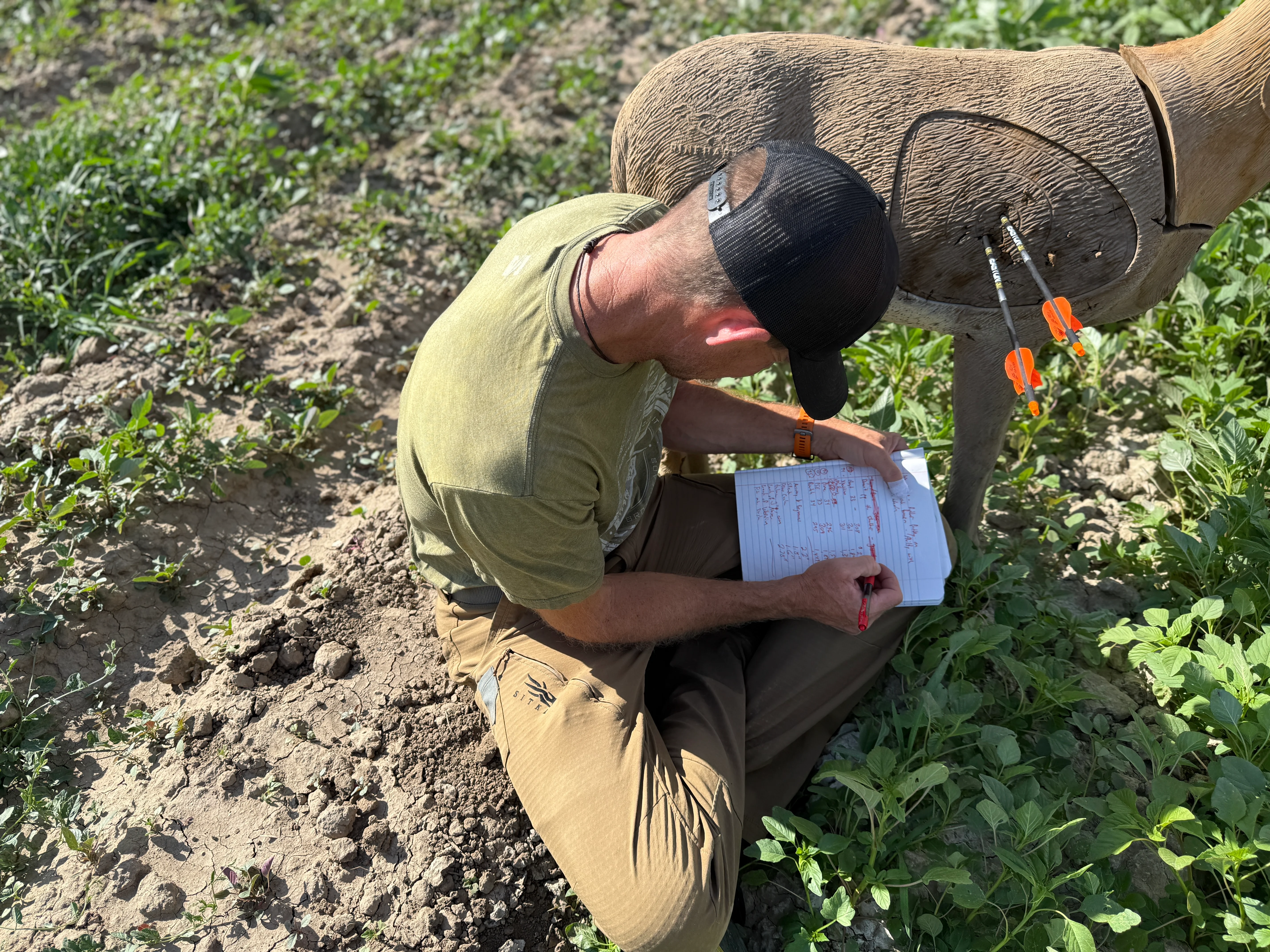A person writes in a notebook while sitting next to a 3D archery deer target.