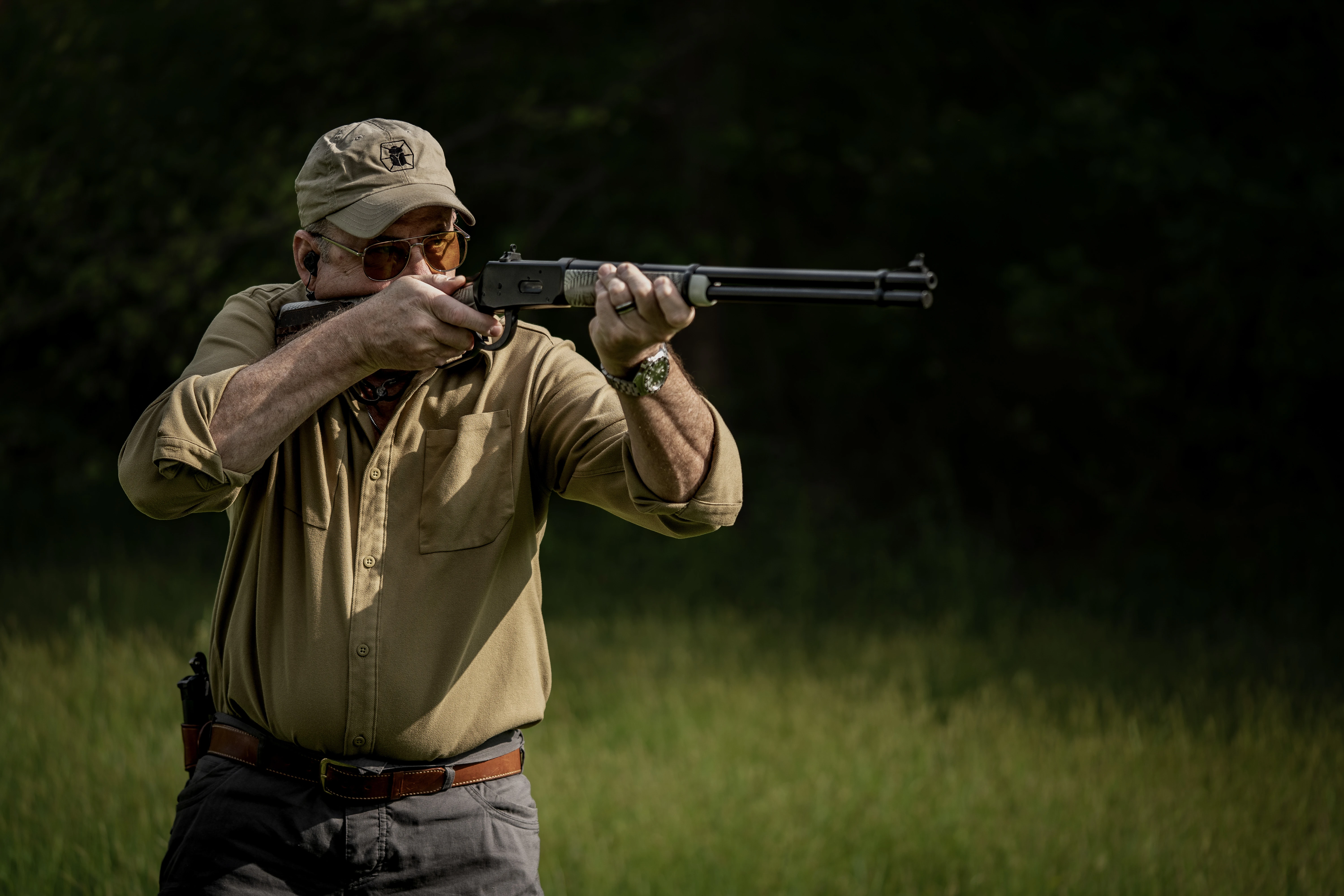 A shooter fires an open-sighted lever-action rifle in a field. 