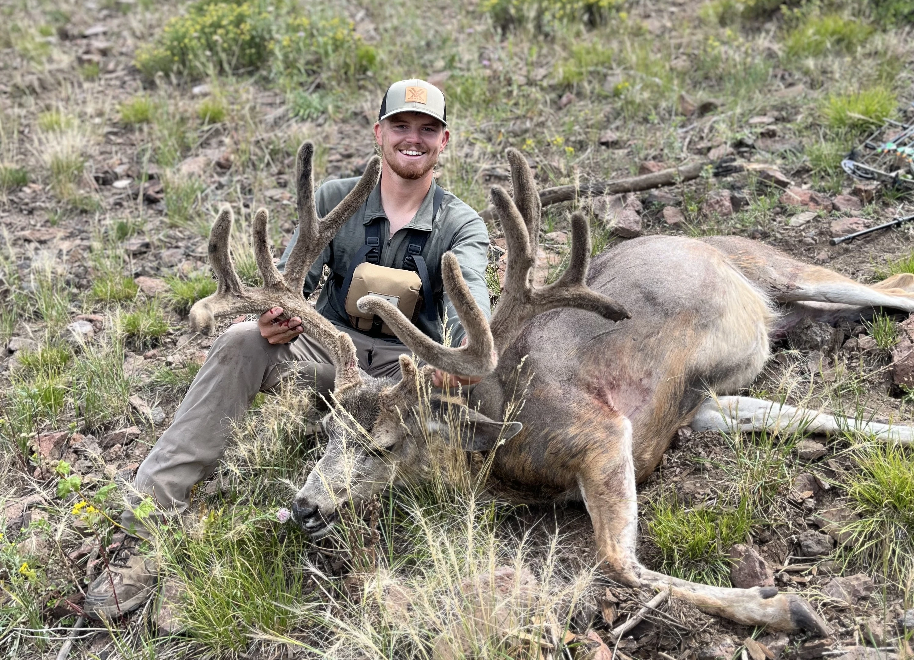 Hunter Keaton Artherton sits on a mountain slope and shows off a huge velvet mule deer he took with bow. 